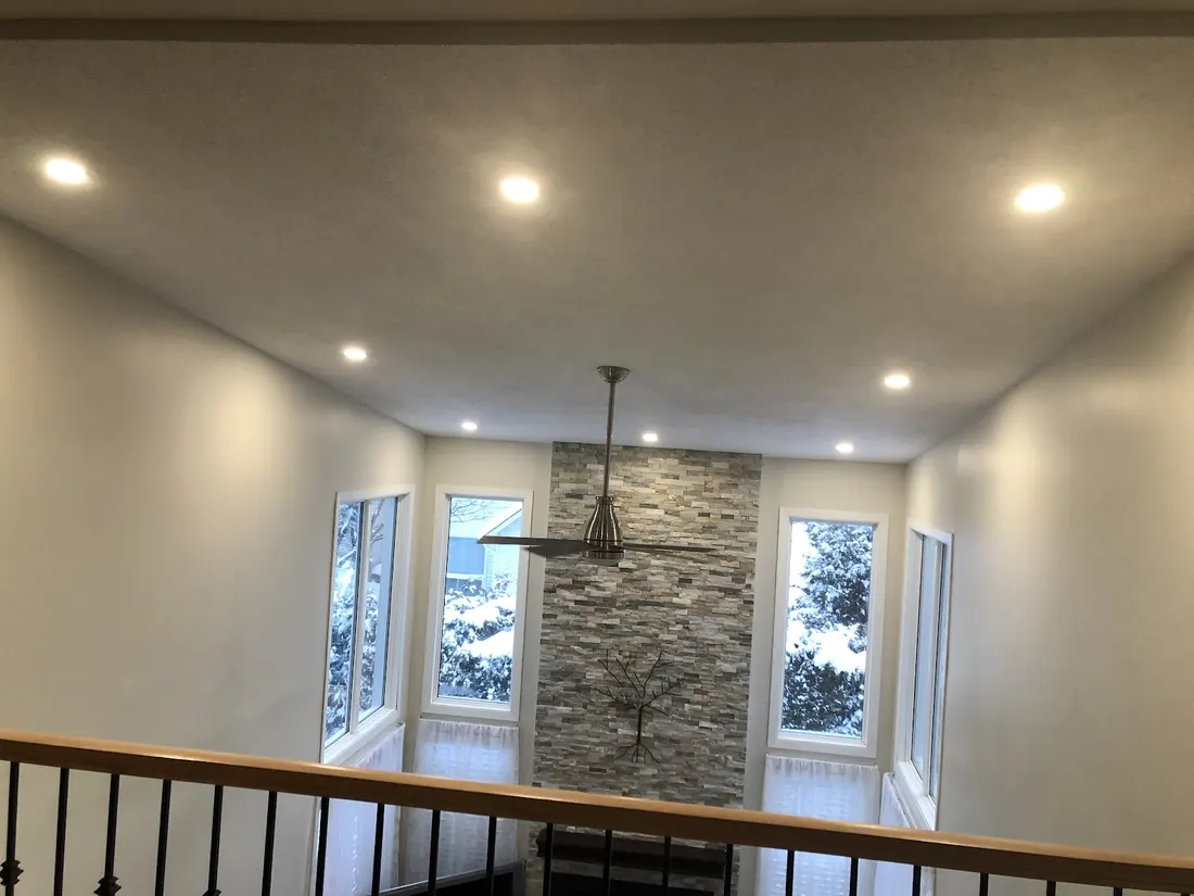 A tall room seen from a railing, with pot lights across the ceiling, a ceiling fan and a stone feature wall between tall windows.